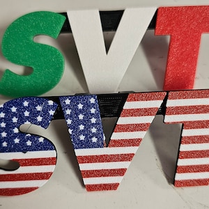 May include: A set of three-dimensional letters spelling "SVT". The top row features green, white, and red letters, while the bottom row displays letters with an American flag design. The letters are mounted on black supports.
