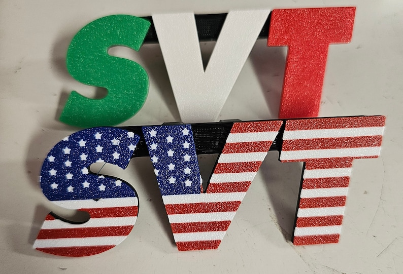 May include: A set of three-dimensional letters spelling "SVT". The top row features green, white, and red letters, while the bottom row displays letters with an American flag design. The letters are mounted on black supports.