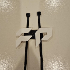 May include: A black and white 3D-printed cable tie holder. The holder features two black cable ties with white geometric shapes attached to a black base. The design is modern and minimalist.