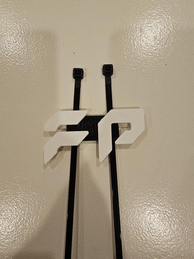 May include: A black and white 3D-printed cable tie holder. The holder features two black cable ties with white geometric shapes attached to a black base. The design is modern and minimalist.