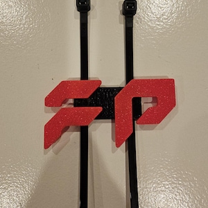May include: A black and red 3D-printed cable tie holder. The holder features a red, glittery logo design and is mounted on two black cable ties. The background is a plain, off-white surface.