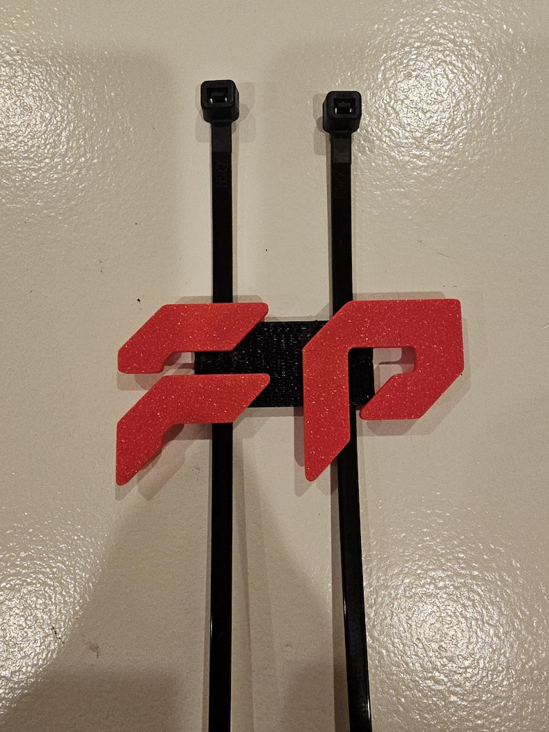 May include: A black and red 3D-printed cable tie holder. The holder features a red, glittery logo design and is mounted on two black cable ties. The background is a plain, off-white surface.