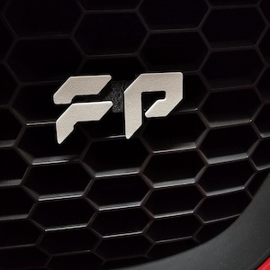 May include: Close-up of a car's front grill with a white "FP" emblem. The emblem is centered on a black honeycomb-patterned grill. The car's red bumper is visible at the bottom of the image.