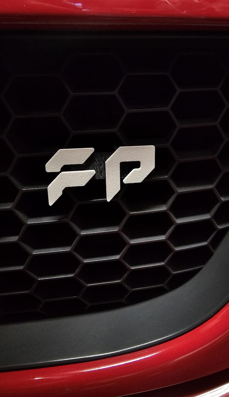 May include: Close-up of a car's front grill with a white "FP" emblem. The emblem is centered on a black honeycomb-patterned grill. The car's red bumper is visible at the bottom of the image.
