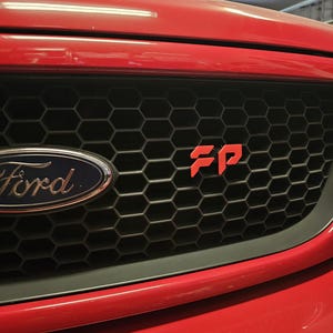 May include: Close-up of a red Ford vehicle's front grille. The black honeycomb grille features a red "FP" emblem and the Ford logo. The vehicle's red bumper and hood are visible.