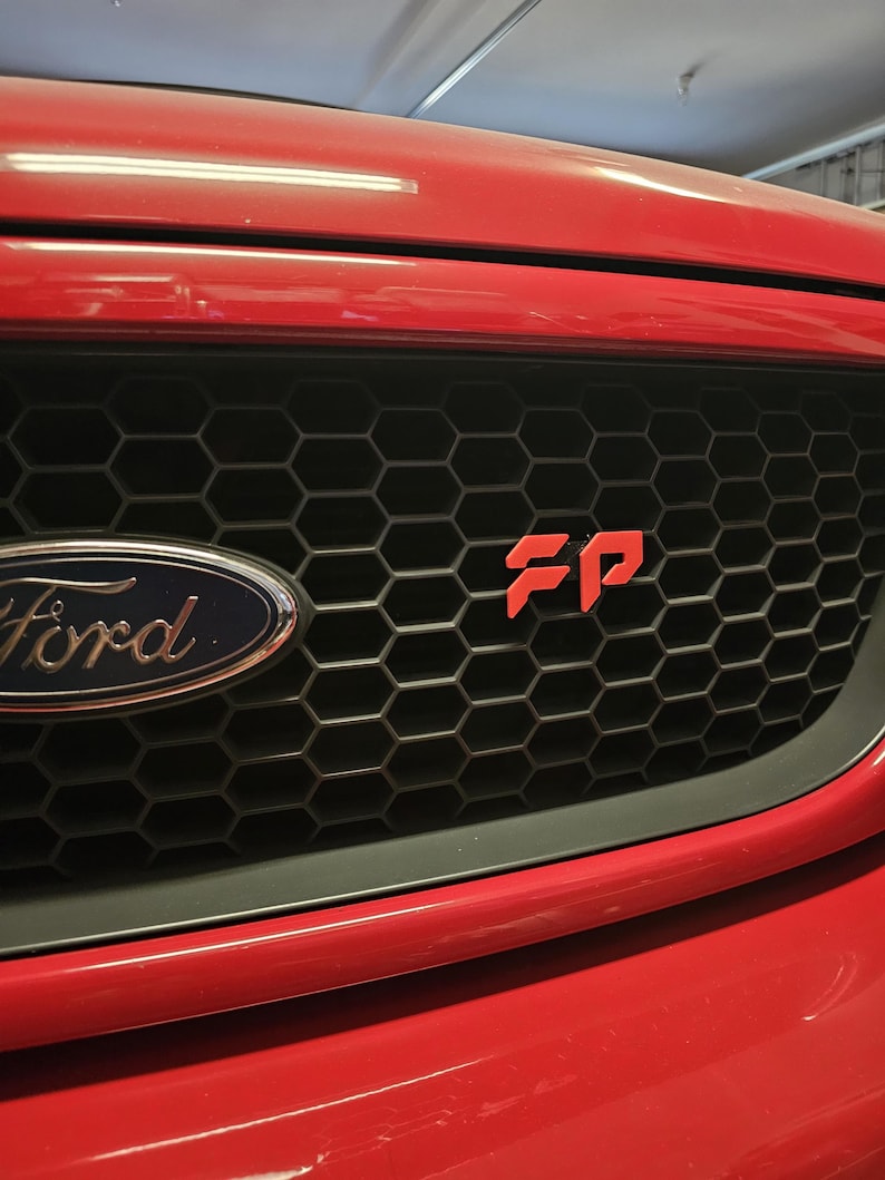 May include: Close-up of a red Ford vehicle's front end, showcasing the black honeycomb grille and the Ford emblem. A red "FP" emblem is prominently displayed on the grille. The car's red bumper is visible.