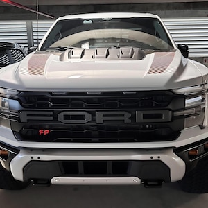 Puede incluir: Un camión Ford Raptor blanco con una parrilla negra y el logotipo de Ford. El capó tiene rayas rojas y blancas. El parachoques delantero es negro y blanco. El camión está estacionado en un garaje.