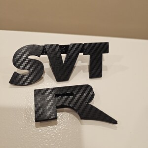 May include: Black carbon fiber SVT R emblem. The letters are separate and have a textured appearance. The emblem is designed to be mounted on a vehicle.