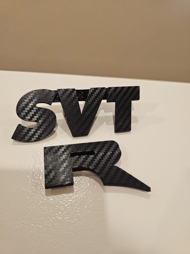 May include: Black carbon fiber SVT R emblem. The letters are separate and have a textured appearance. The emblem is designed to be mounted on a vehicle.