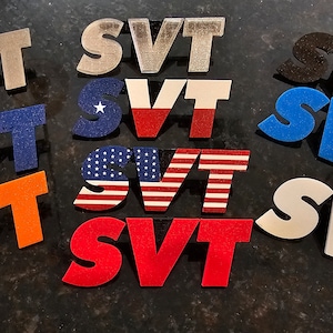May include: A collection of various SVT logo designs in different colors and patterns. The logos include solid colors like orange, red, and blue, as well as designs featuring the American and Texas flags. The logos are displayed on a dark surface.