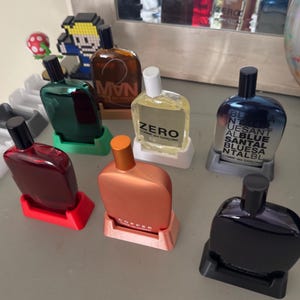 Puede incluir: Una colección de ocho botellas de perfume en varios colores, incluyendo rojo, verde, marrón y negro, se muestran en soportes individuales. Las botellas están etiquetadas con los nombres "Man", "Zero", "Blunt" y "Copper".