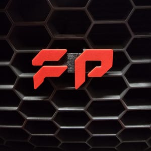 May include: Close-up of a black honeycomb grille with a bright red FP emblem. The emblem is a stylized design, likely a logo or badge, and stands out against the dark background. The image is well-lit, highlighting the texture of the grille.