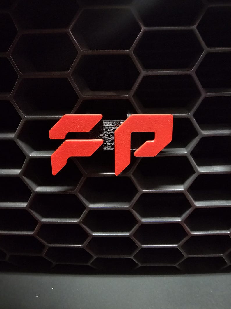 May include: Close-up of a black honeycomb grille with a bright red FP emblem. The emblem is a stylized design, likely a logo or badge, and stands out against the dark background. The image is well-lit, highlighting the texture of the grille.