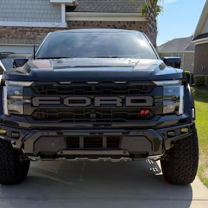 May include: A black Ford Raptor truck, viewed head-on, showcasing the prominent FORD lettering across the grille. The truck has large, rugged tires and a black front bumper. The image is taken outdoors in front of a house.
