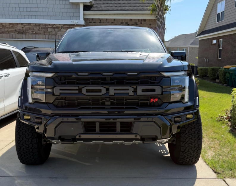 May include: A black Ford Raptor truck, viewed head-on, showcasing the prominent FORD lettering across the grille. The truck has large, rugged tires and a black front bumper. The image is taken outdoors in front of a house.