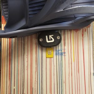 Puede incluir: Tabla de snowboard con un diseño de rayas verticales coloridas. Una fijación gris oscuro en la parte superior, con una pieza negra de forma ovalada con el logotipo "LS" blanco. Una etiqueta amarilla con el mismo logotipo debajo. La palabra "BURTON" en la parte inferior.