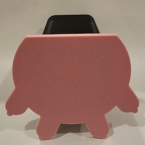 Puede incluir: Un objeto de espuma rosa texturizada con forma de cerdo, con cuatro patas pequeñas y un cuerpo redondeado. El objeto con forma de cerdo está sobre un fondo blanco, con un objeto negro parcialmente visible detrás.