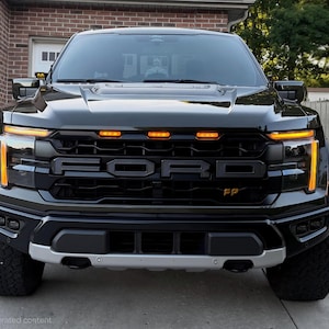 May include: Front view of a black Ford Raptor truck. The truck has a black grille with the Ford logo, amber running lights, and a silver front bumper. The tires are large and black.