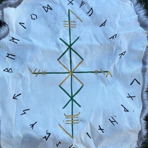 May include: A white hide rug with a grey fur trim. The rug features a central geometric design in green and gold, surrounded by black runic symbols. The rug is displayed on green grass.