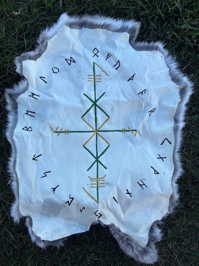 May include: A white hide rug with a grey fur trim. The rug features a central geometric design in green and gold, surrounded by black runic symbols. The rug is displayed on green grass.