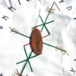 May include: A wooden, oval-shaped rune stone with green markings rests on a white surface adorned with green and brown geometric designs and black runic symbols. The background is a white, textured material.