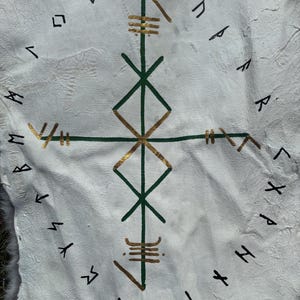 May include: White leather hide featuring a central green and gold geometric design, encircled by black runic symbols. The design includes a cross-like structure with diamond shapes and horizontal lines. The hide has a textured surface, possibly for decorative or ritual use.