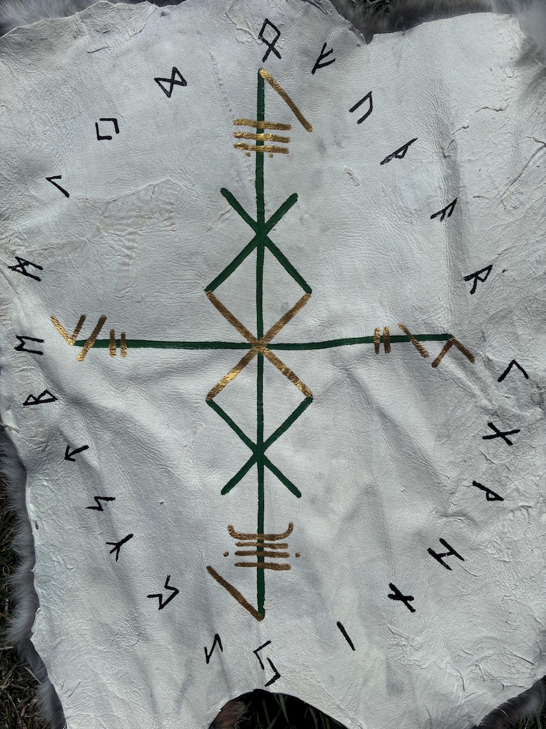 May include: White leather hide featuring a central green and gold geometric design, encircled by black runic symbols. The design includes a cross-like structure with diamond shapes and horizontal lines. The hide has a textured surface, possibly for decorative or ritual use.