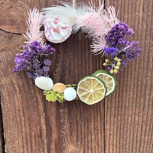 May include: A small, circular wreath featuring a seashell, dried lime slices, and purple flowers. The wreath is adorned with pink feathers and small white and tan stones. It hangs from a natural twine string against a rustic, wooden background.