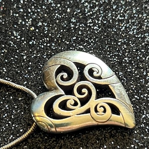 May include: A silver-toned heart pendant with an openwork design featuring swirling patterns. The pendant hangs from a thin silver chain. The background is a dark, glittery surface.