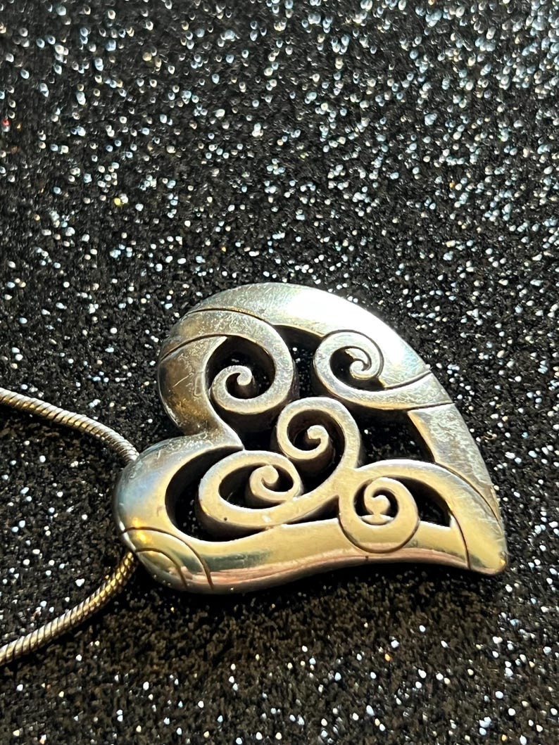 May include: A silver-toned heart pendant with an openwork design featuring swirling patterns. The pendant hangs from a thin silver chain. The background is a dark, glittery surface.
