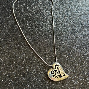 May include: Silver-toned necklace featuring a heart-shaped pendant with an openwork design. The pendant hangs from a delicate chain with a lobster clasp closure. The heart has a swirling pattern within its outline.