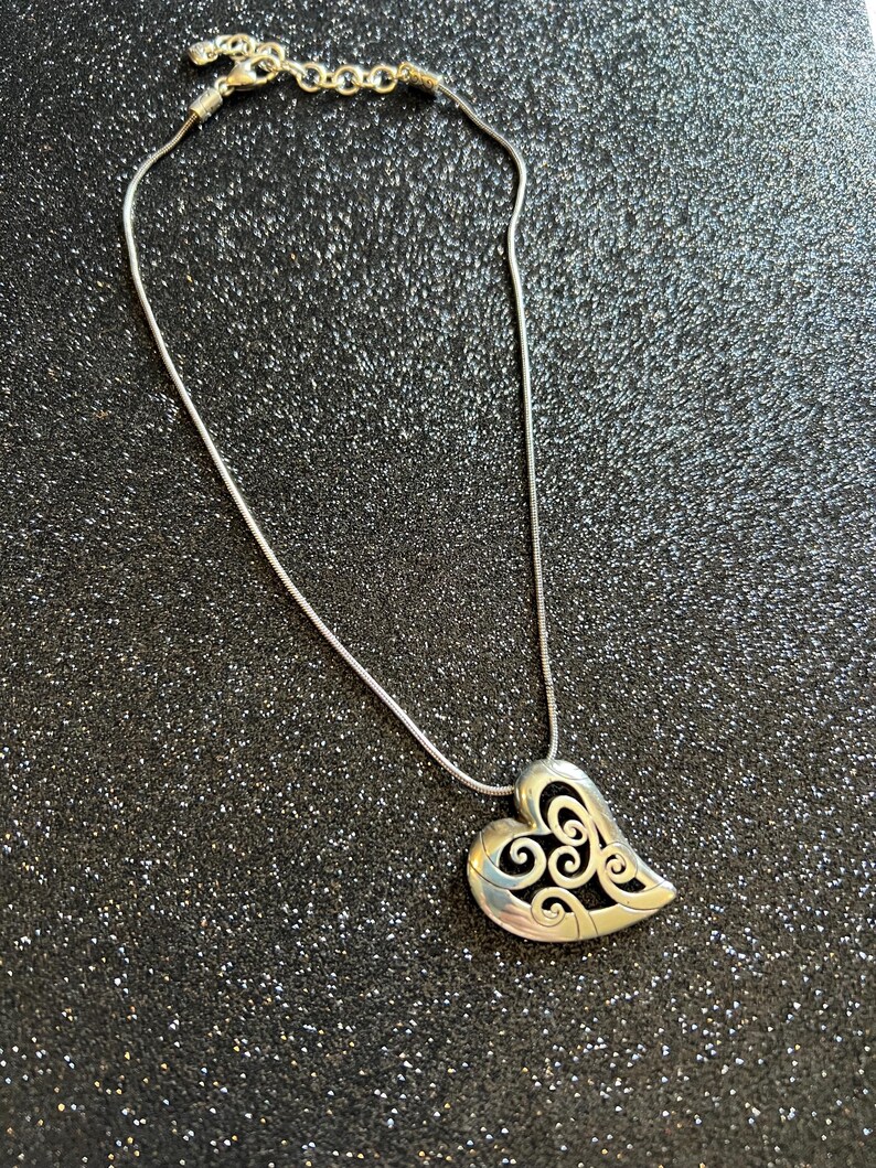 May include: Silver-toned necklace featuring a heart-shaped pendant with an openwork design. The pendant hangs from a delicate chain with a lobster clasp closure. The heart has a swirling pattern within its outline.