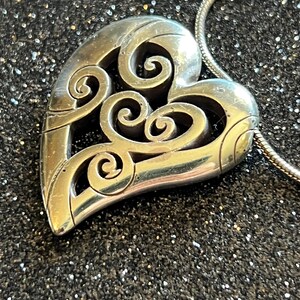 May include: Silver-toned heart pendant with intricate scrollwork design. The pendant is suspended from a silver chain. The heart shape is detailed with openwork, creating a delicate and elegant aesthetic. The background is a dark, glittery surface.