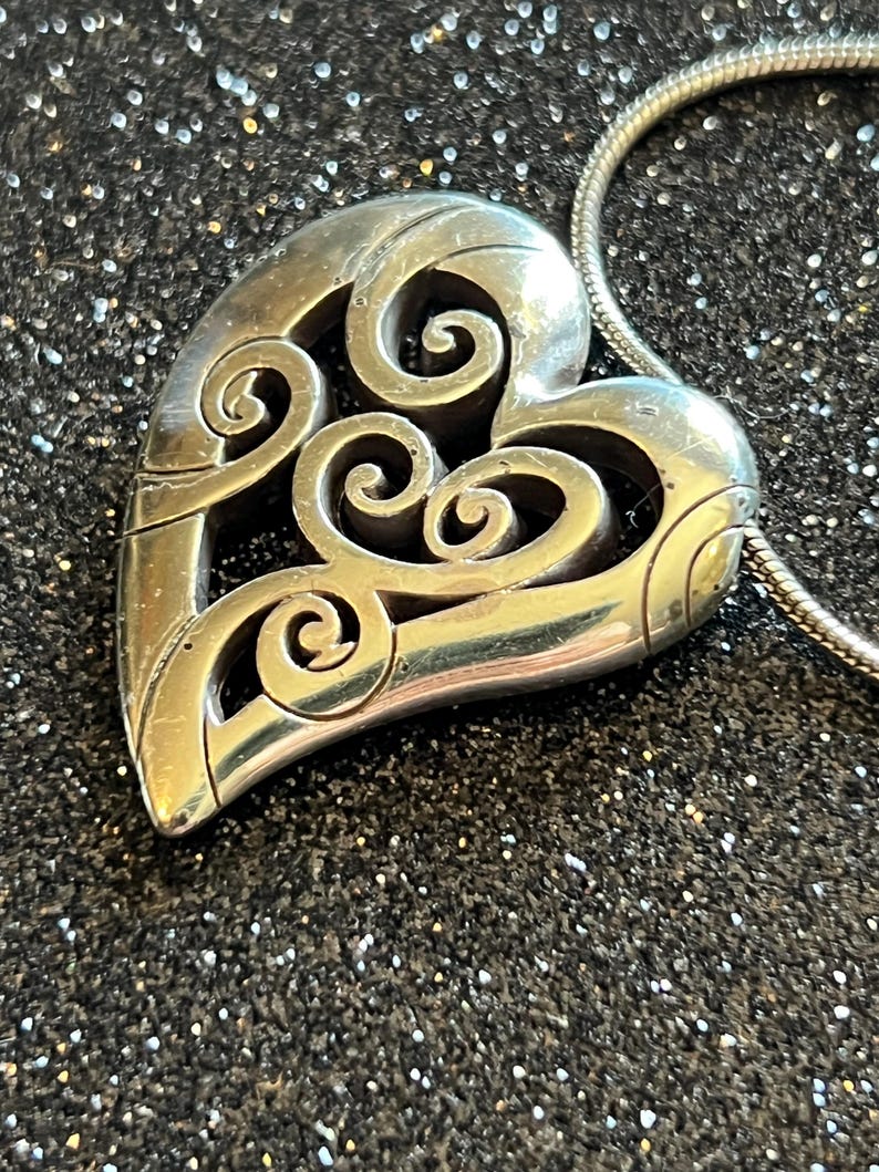 May include: Silver-toned heart pendant with intricate scrollwork design. The pendant is suspended from a silver chain. The heart shape is detailed with openwork, creating a delicate and elegant aesthetic. The background is a dark, glittery surface.