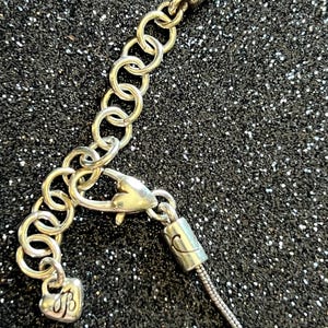 May include: Silver-toned necklace with a heart-shaped charm and a lobster clasp. The necklace features a series of small, linked rings for adjustable sizing. The charm has an engraved initial. The background is a dark, glittery surface.