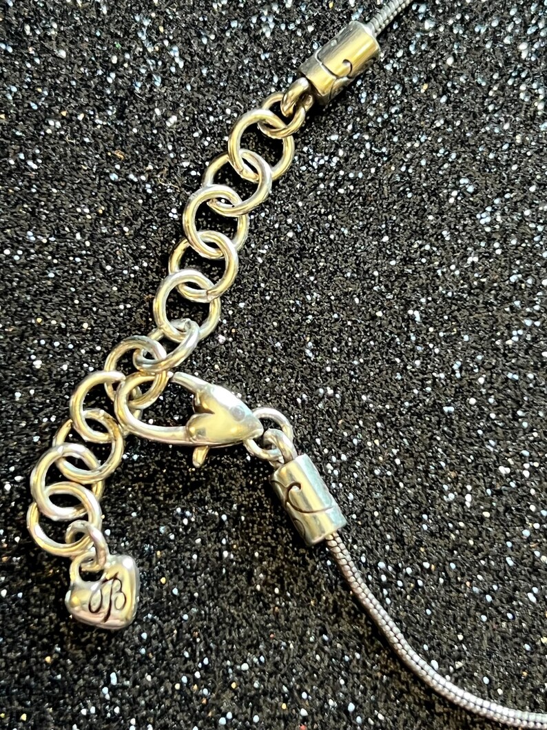 May include: Silver-toned necklace with a heart-shaped charm and a lobster clasp. The necklace features a series of small, linked rings for adjustable sizing. The charm has an engraved initial. The background is a dark, glittery surface.