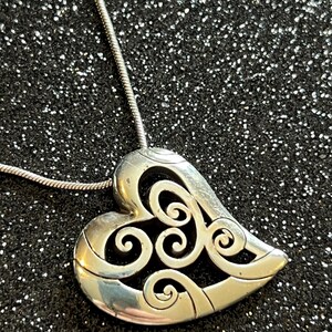 May include: Silver-toned heart pendant necklace with an intricate openwork design. The heart-shaped pendant features swirling patterns and is suspended from a delicate silver chain. The necklace is displayed against a dark, glittery background.