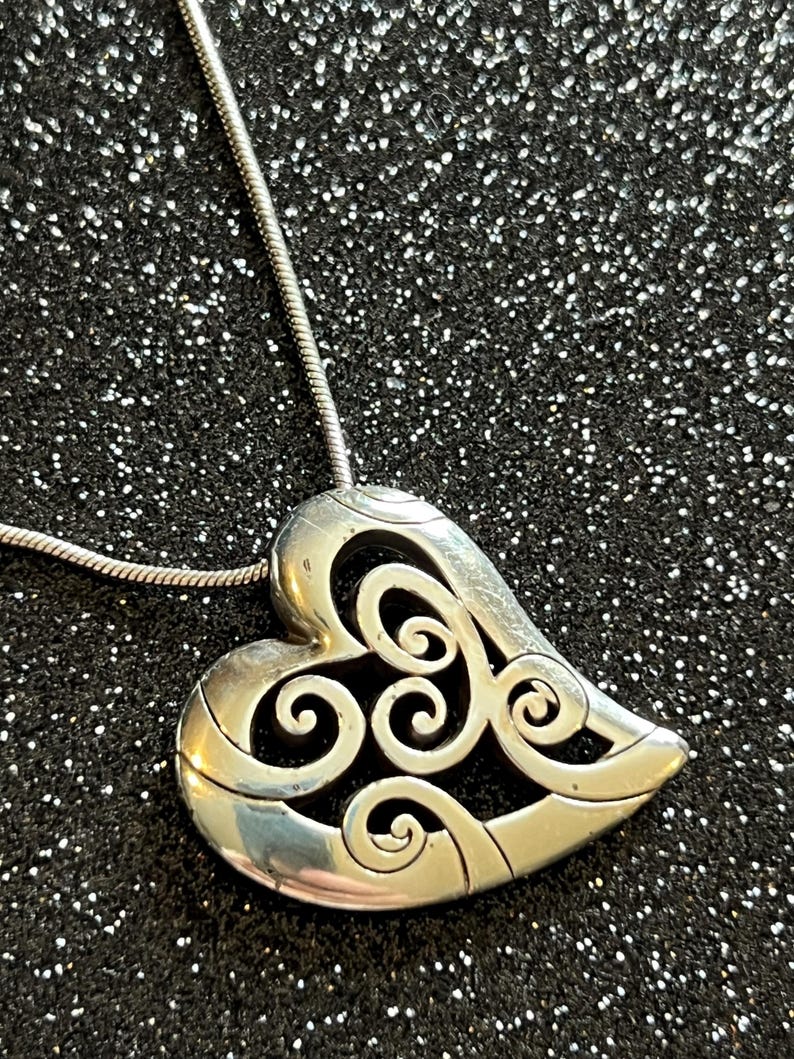 May include: Silver-toned heart pendant necklace with an intricate openwork design. The heart-shaped pendant features swirling patterns and is suspended from a delicate silver chain. The necklace is displayed against a dark, glittery background.