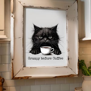 Puede incluir: Impresión enmarcada que presenta un gato negro gruñón con una taza de café. El gato tiene una expresión severa y está posicionado detrás de una taza blanca. El texto "Grumpy before Coffee" está debajo del gato. El marco es blanco con un acabado envejecido.
