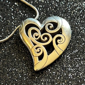 May include: Silver-toned heart pendant necklace with an intricate openwork design. The heart features swirling patterns and is suspended from a delicate chain. The necklace is displayed against a dark, glittery background.