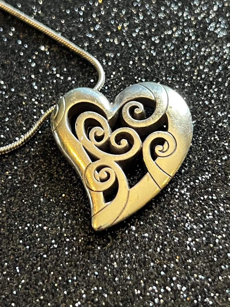 May include: Silver-toned heart pendant necklace with an intricate openwork design. The heart features swirling patterns and is suspended from a delicate chain. The necklace is displayed against a dark, glittery background.