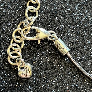 May include: Close-up of a silver-toned necklace clasp and chain. The clasp is heart-shaped, and the chain is made of small, interconnected rings. The background is a black, glittery surface.