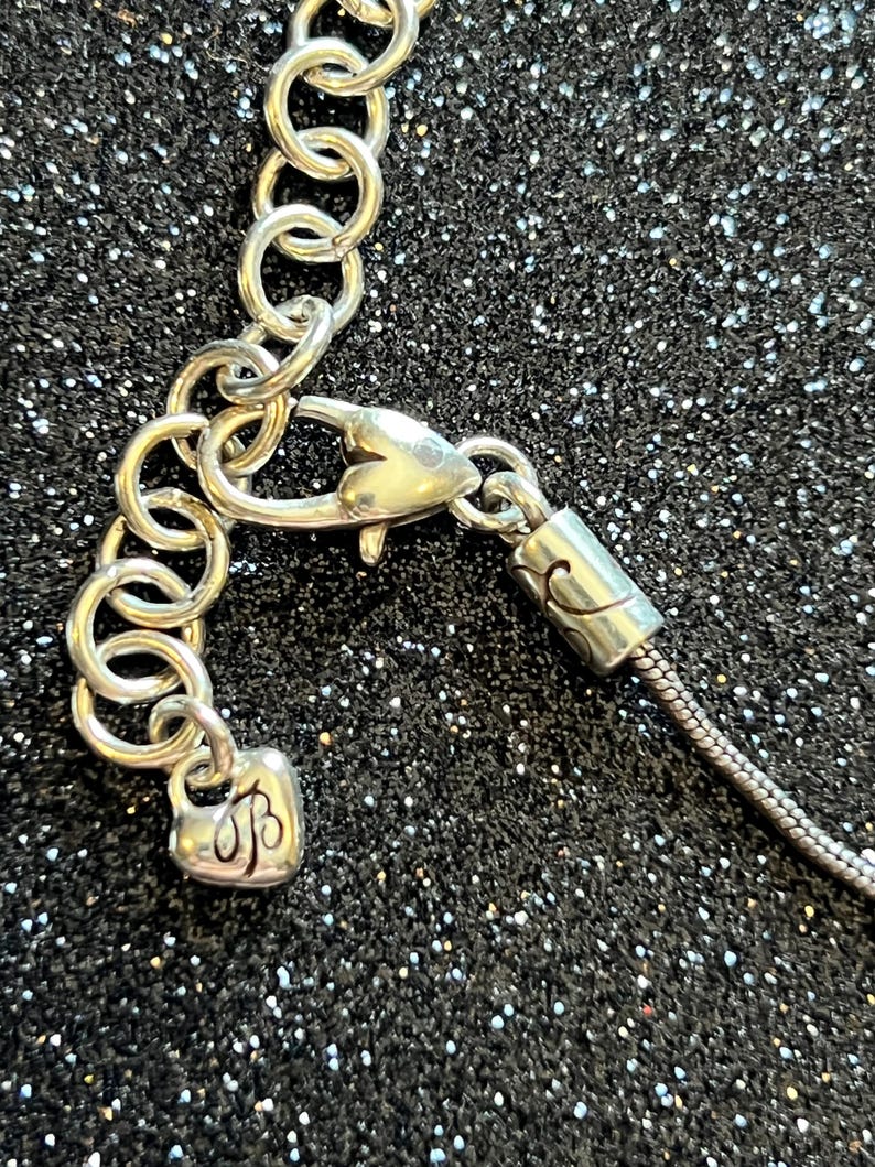 May include: Close-up of a silver-toned necklace clasp and chain. The clasp is heart-shaped, and the chain is made of small, interconnected rings. The background is a black, glittery surface.