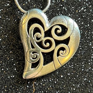 May include: Silver-toned heart pendant with an openwork design of swirling patterns. The pendant hangs from a thin, silver chain. The heart shape is detailed with a textured finish, creating a vintage or antique aesthetic. The background is a dark, glittery surface.