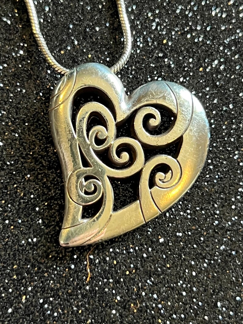 May include: Silver-toned heart pendant with an openwork design of swirling patterns. The pendant hangs from a thin, silver chain. The heart shape is detailed with a textured finish, creating a vintage or antique aesthetic. The background is a dark, glittery surface.