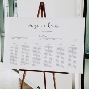May include: A white wedding seating chart with the names Megan and Kevin, the date April 28, 2022, and the location London. The chart is on an easel with a wooden frame.