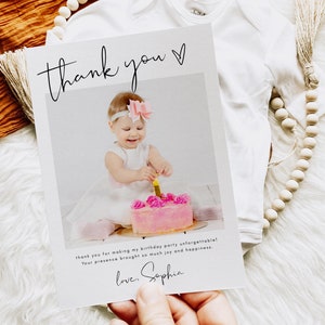 May include: A thank you card with a photo of a baby girl at her first birthday party. The card has the words "thank you" in script, and a message thanking guests. A white onesie and wooden beads are in the background.