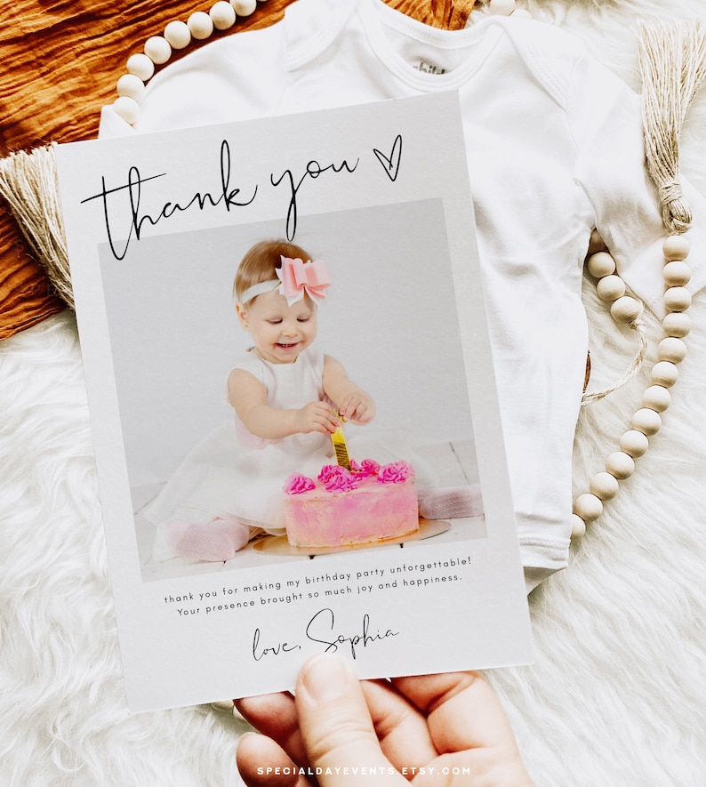 May include: A thank you card with a photo of a baby girl at her first birthday party. The card has the words "thank you" in script, and a message thanking guests. A white onesie and wooden beads are in the background.