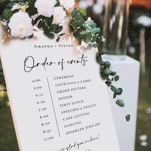 May include: A wedding event schedule with the names "AMANDA + STEVEN" at the top. The schedule lists the order of events, including the ceremony, dinner, first dance, and cake cutting. The sign is decorated with white roses and greenery.