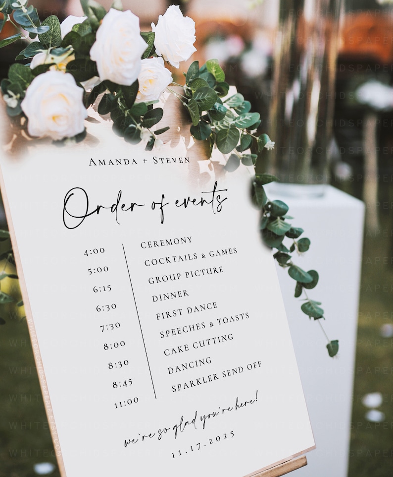 May include: A wedding event schedule with the names "AMANDA + STEVEN" at the top. The schedule lists the order of events, including the ceremony, dinner, first dance, and cake cutting. The sign is decorated with white roses and greenery.